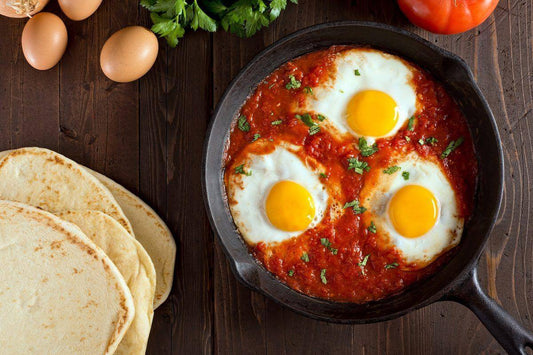 Three eggs cooked in a spicy tomato sauce, served in a skillet with tortillas and fresh ingredients on the side.
