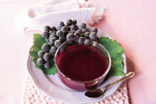Bowl of Vino Cotto syrup surrounded by fresh black grapes and green leaves on a decorative plate.