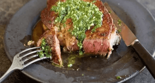 Juicy steak topped with green sauce on a black plate with a fork and knife beside it.