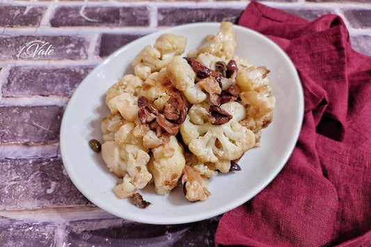 Plate of cooked cauliflower with capers and olives served on a white dish, alongside a burgundy napkin.
