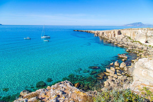 Crystal clear waters and rocky coastline at Favignana, Isole Egadi, with boats anchored in the tranquil bay.