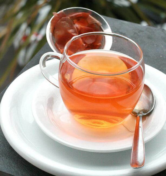 A clear glass cup filled with amber tea on a white saucer, accompanied by a spoon and tea leaves.