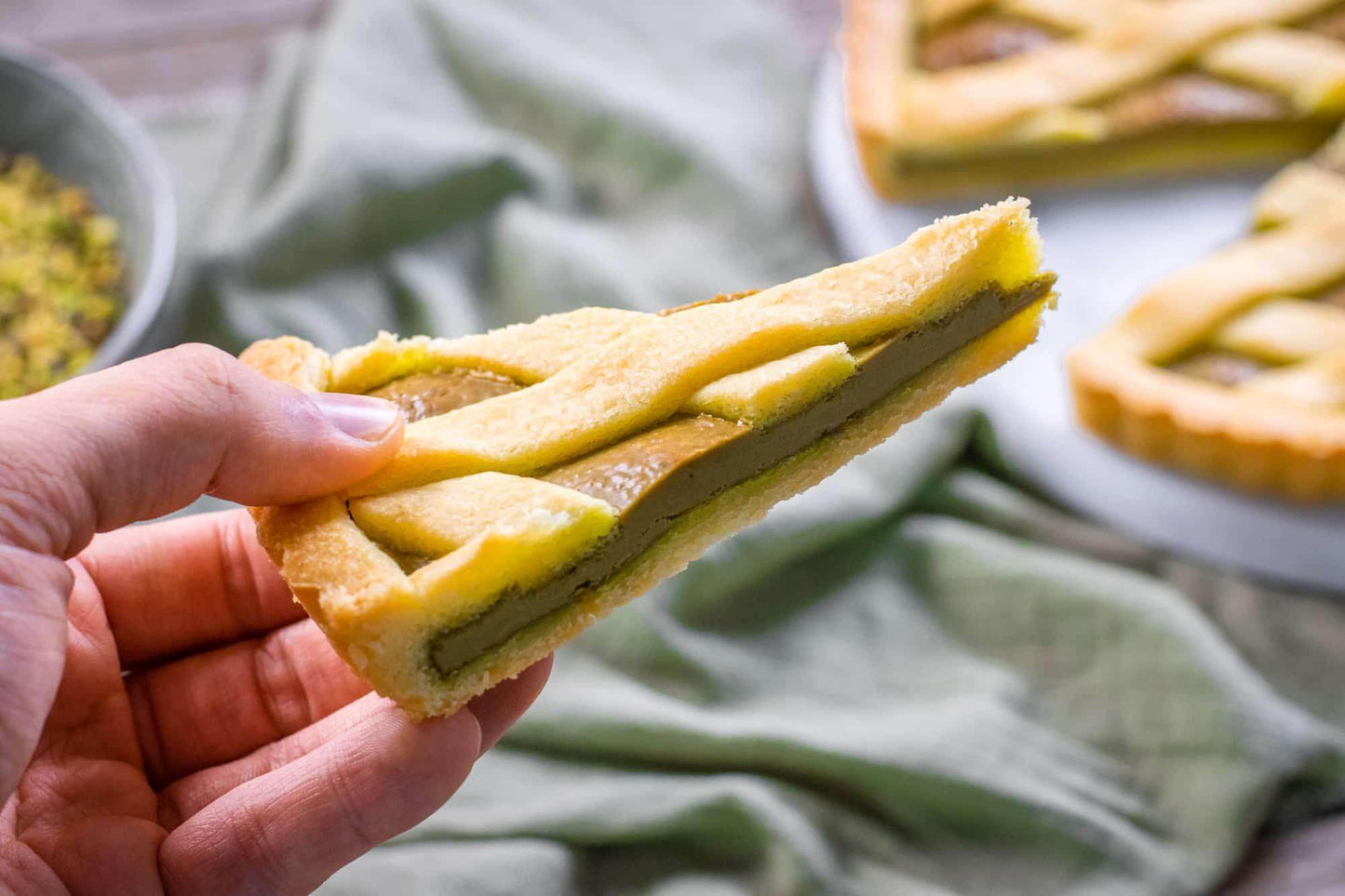 Slice of handcrafted Sicilian pistachio crostata held by hand, showcasing its rich filling and golden crust.