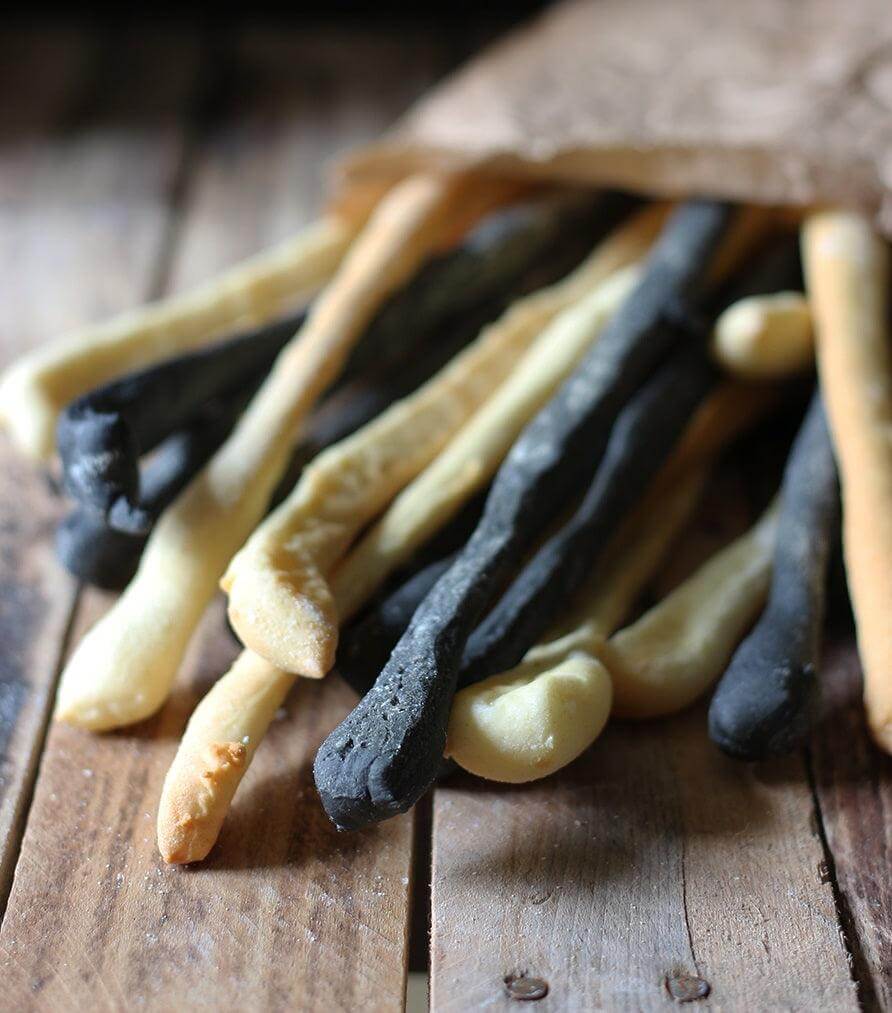 Artisanal charcoal grissini sticks in various colors, served in a rustic paper bag on a wooden table.