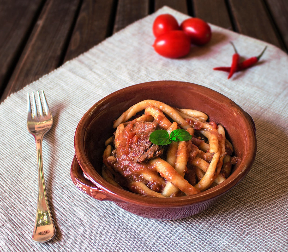Ragù di maiale pronto alla calabrese servito con pasta in una ciotola di terracotta, decorato con pomodorini e peperoncino.