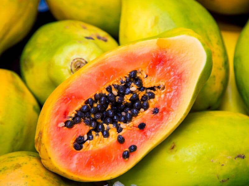Freshly cut papaya with vibrant orange flesh and black seeds, surrounded by whole green papayas.