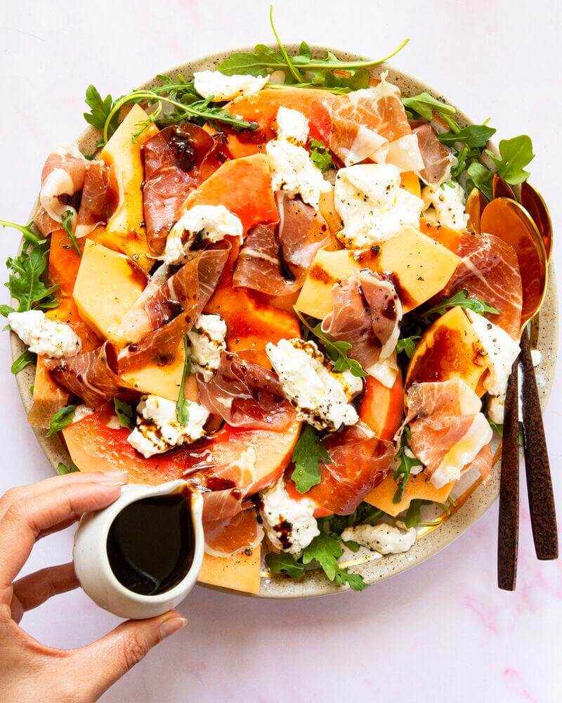 Fresh salad with melon, prosciutto, arugula, and creamy cheese, drizzled with balsamic dressing.