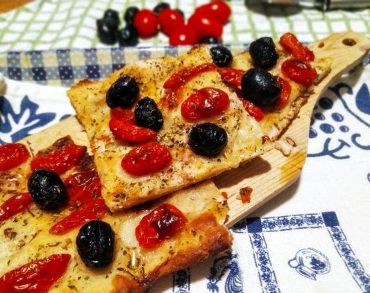 Focaccia Mediterranea con pomodorini rossi e olive nere su un tagliere di legno.