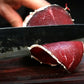 Salame di Capra being sliced with a knife, showcasing its rich red color and texture.