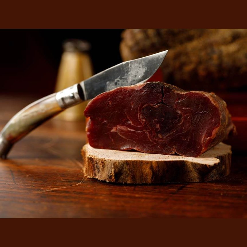 Salame di Capra trancio c.a 500 gr. su un tagliere di legno con un coltello in acciaio a lato.