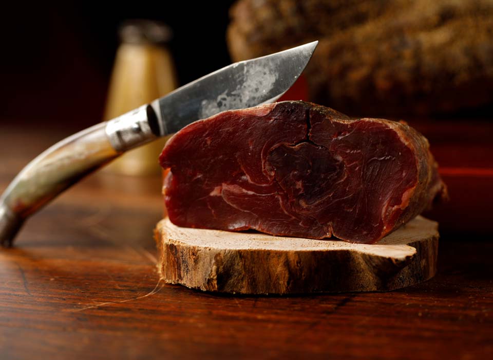 Salame di Capra trancio c.a 500 gr. su un tagliere di legno con un coltello in acciaio a lato.